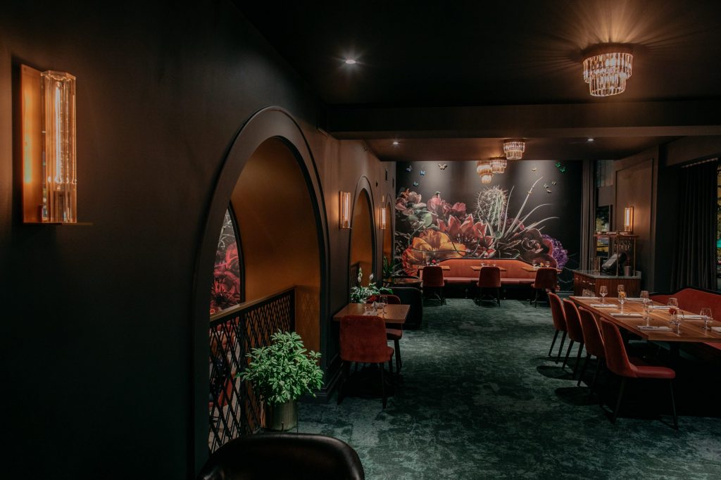 The interior of Monarch Prime & Bar is lush and moody with plush velvet carpeting and chairs. A large floral mural is on the far wall, and dark archways on the left are punctuated with glass light fixtures that throw golden glows around the room.
